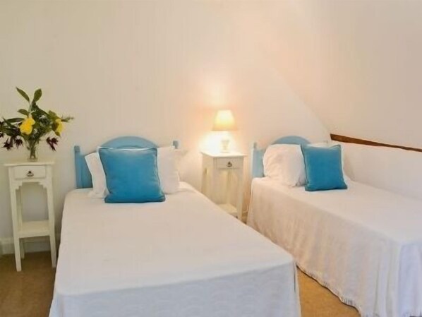 Habitación de hotel con dos camas individuales, cada una con almohada azul y ropa de cama blanca. Hay una mesita de noche pequeña con una lámpara y un jarrón con flores.