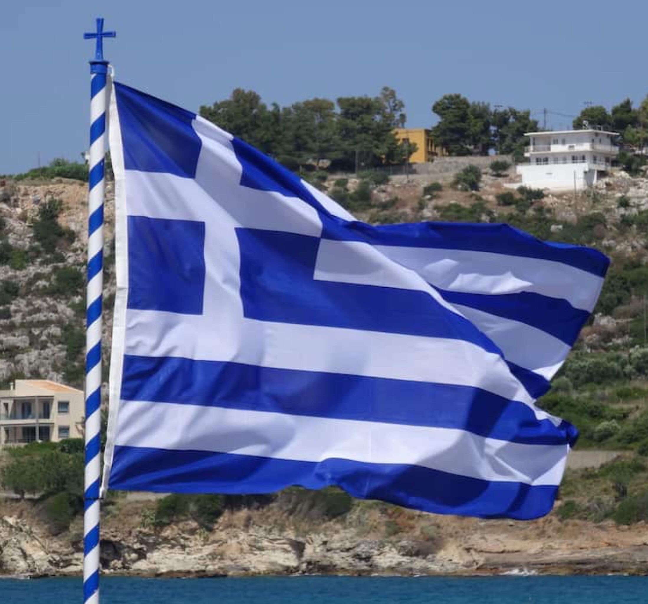 A flag on a flagpole by a coastal area with buildings in the background.