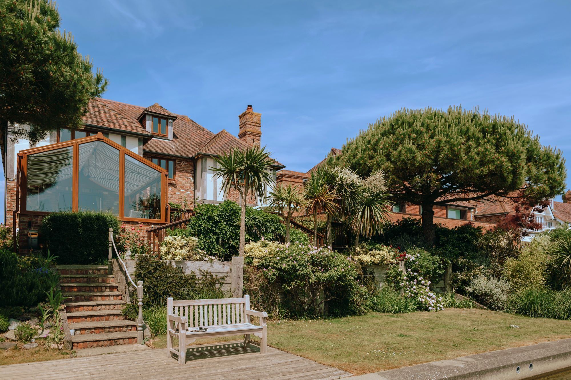 A house with a large glass conservatory, a wooden deck, and a well-maintained garden with various plants and a bench.