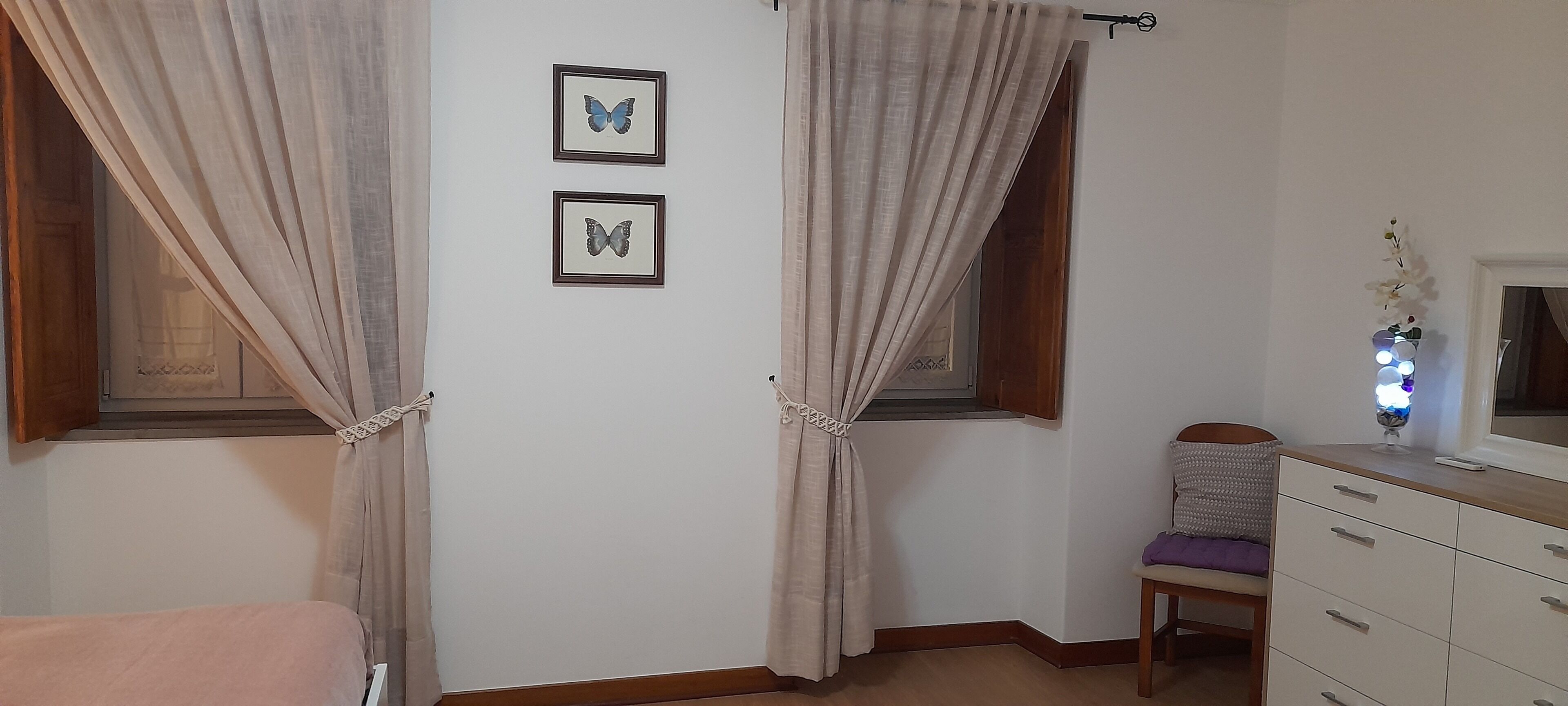 Ein Schlafzimmer mit zwei Fenstern, einem Bett, einer Kommode und zwei gerahmten Schmetterlingsbildern an der Wand.