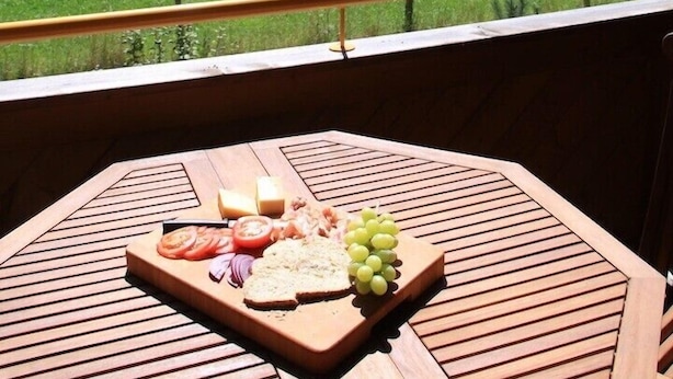 Un tavolo di legno con un tagliere, uva e pomodori su un balcone con vista su un campo verde e montagne.