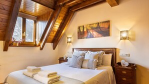 Een slaapkamer met een groot bed, houten balken in het plafond, een schilderij van een bos en een nachtkastje met een lamp.