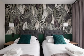 A hotel room with two beds, each with a white bedspread and teal pillows, against a wall with a leaf-pattern wallpaper.