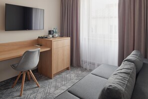 A modern hotel room with a wooden desk, a grey chair, a sofa, and a TV mounted on the wall.