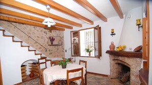 Zona pranzo con pavimento in pietra, sedie di legno, tavolo con tovaglia bianca, camino e una scala con parete decorata.