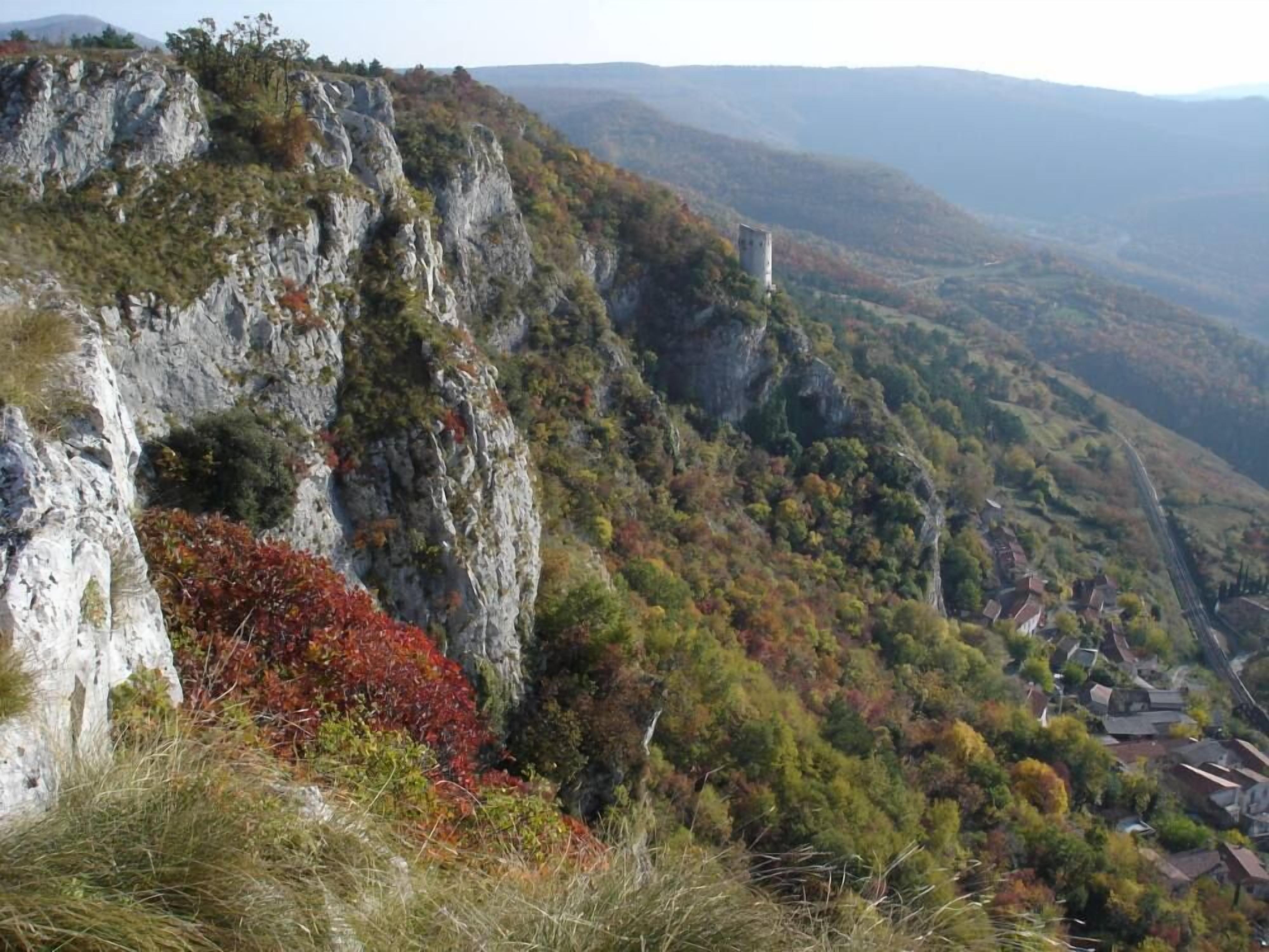 A mountainous landscape with a castle-like structure on a cliff, surrounded by autumn-colored trees and a valley below.