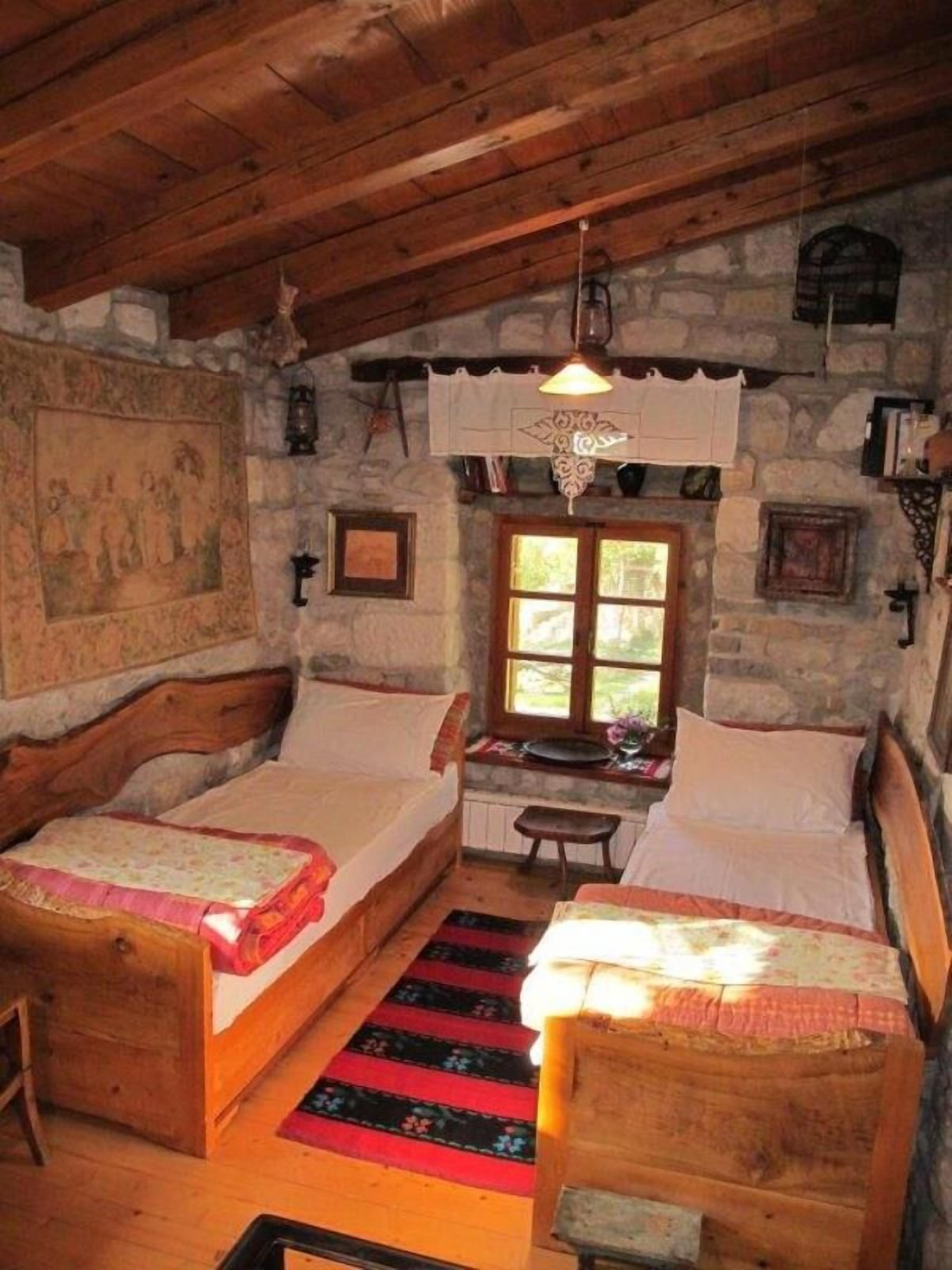 A rustic bedroom with two wooden beds, a stone wall, a window, and a hanging light fixture.