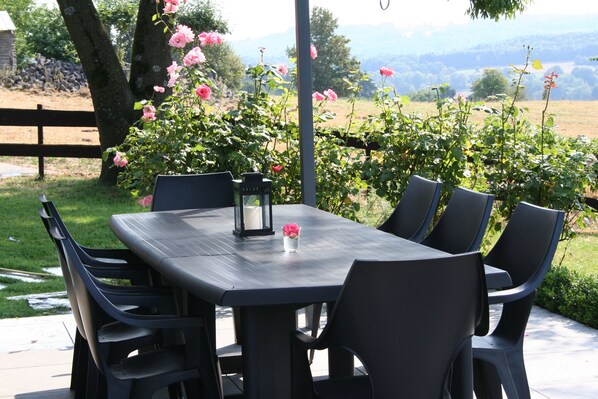 An outdoor patio with a dark wooden table and six chairs, surrounded by lush greenery and pink flowers. - Magnificent character cottage in the heart of the Ardennes (Stavelot)
