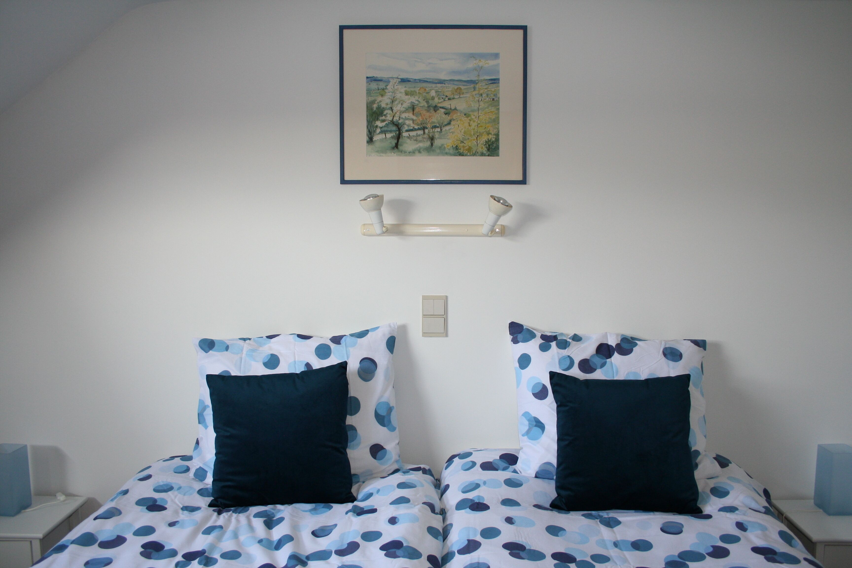 A bedroom with two beds, each with a white and blue polka dot bedspread and matching pillows. There is a framed painting above the beds and a small shelf below.