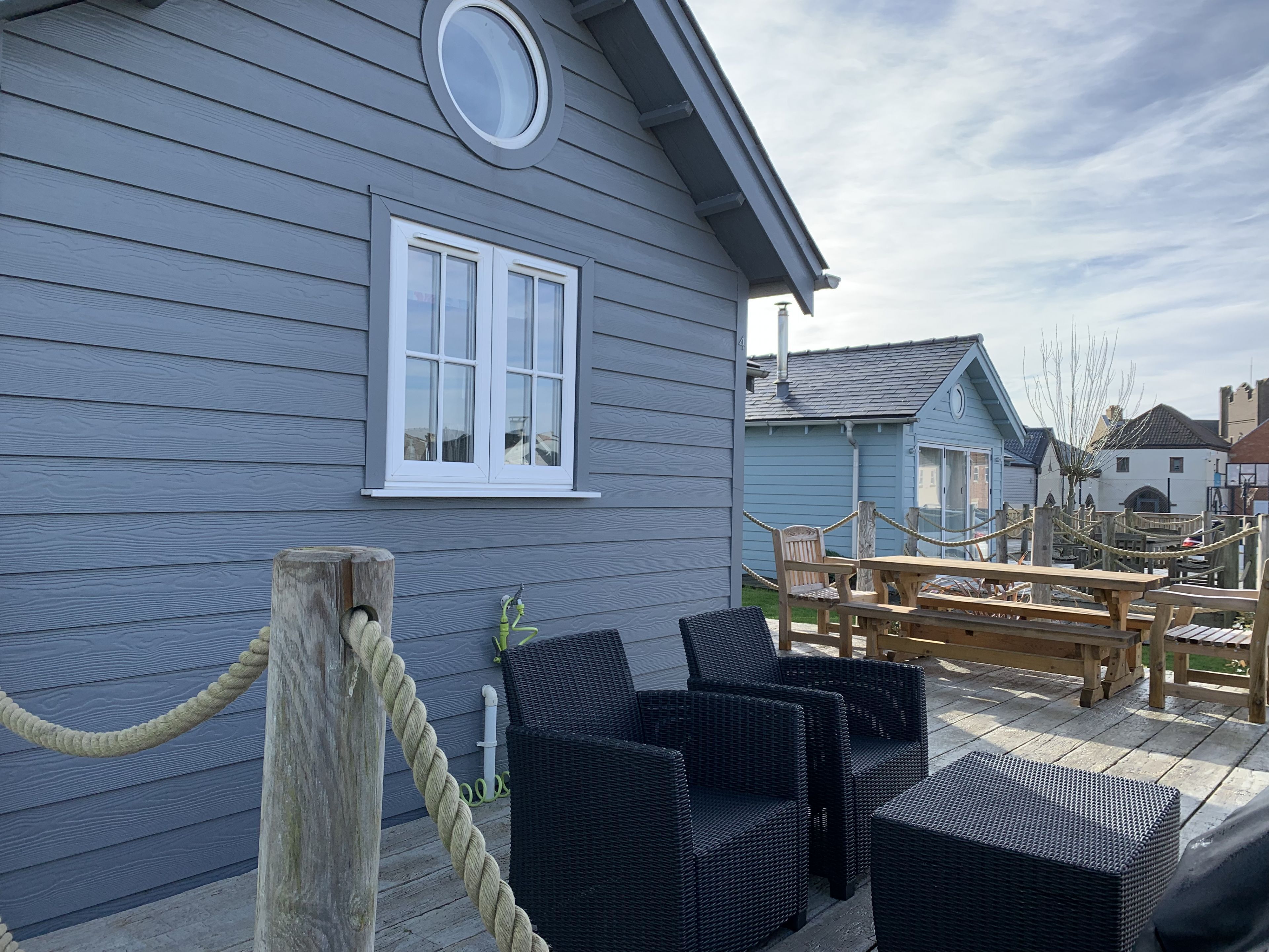 A wooden deck with wicker chairs and a table, a rope barrier, and a building with a window and a circular skylight.