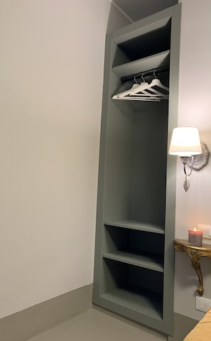 Un placard haut et étroit, doté de plusieurs étagères et d’une tringle pour suspendre les vêtements, une lampe fixée au mur et une petite table sur laquelle est posée une bougie.