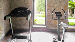 Un gimnasio con cintas de correr y una máquina elíptica, paredes de piedra y una puerta que da a un jardín.