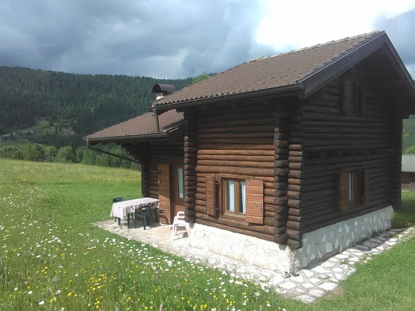 A wooden log cabin with a stone foundation, a small patio with a table and chairs, and a view of a forested valley. - Chalet surrounded by greenery near MTB routes (Castello Tesino)