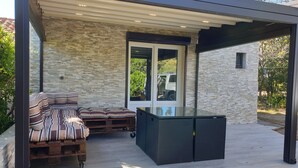 A covered outdoor seating area with a stone wall, a glass door, and a modern outdoor kitchen setup. - house / villa - 1 room - 4 persons (Porto-Vecchio)