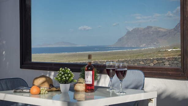 Une table avec une bouteille de vin, deux verres, une orange et un chapeau. Une fenêtre donne sur un paysage côtier.