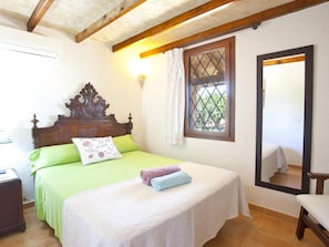 A bedroom with a wooden headboard, a bed with green bedding, a white bedside table, and a window with a view of greenery. - Villa Son Fullos ·Villa with private pool in Santa Margalida (Santa Margalida)