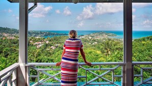 A woman standing on a balcony overlooking a coastal landscape with a view of the sea and a town.
