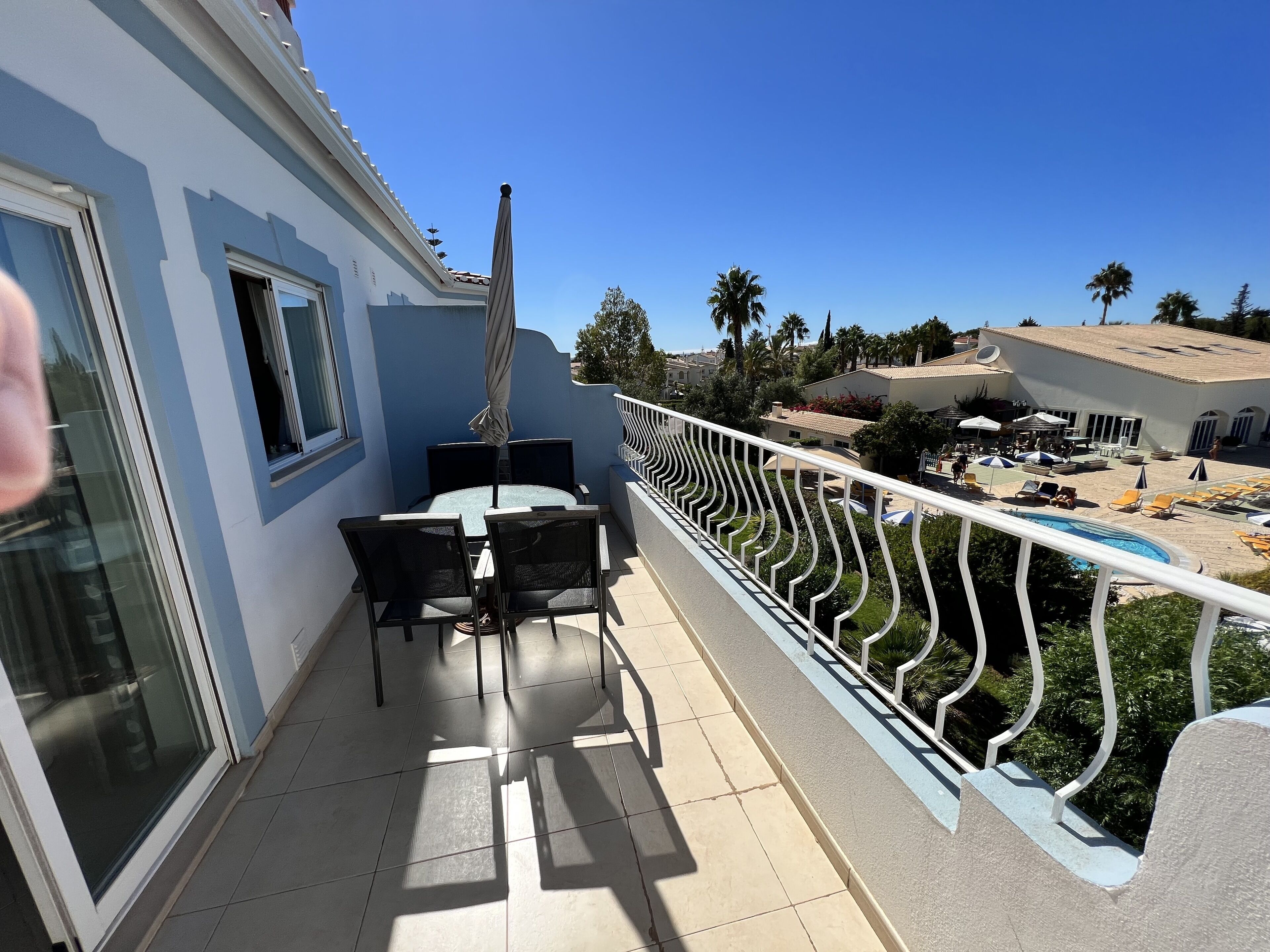 A balcony with a table and chairs, a pool area with lounge chairs, and a view of neighboring buildings and palm trees.