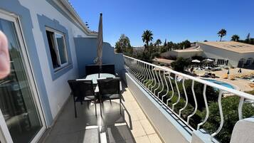 A balcony with a table and chairs, a pool area with lounge chairs, and a view of neighboring buildings and palm trees.