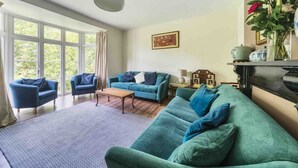 A living room with two sofas, a coffee table, and a fireplace. There is a large window with curtains, a painting on the wall, and a vase with flowers on a shelf. - Large Period House set in Ancient Woodland in Exmoor National Park near Lynmouth (Lynton)