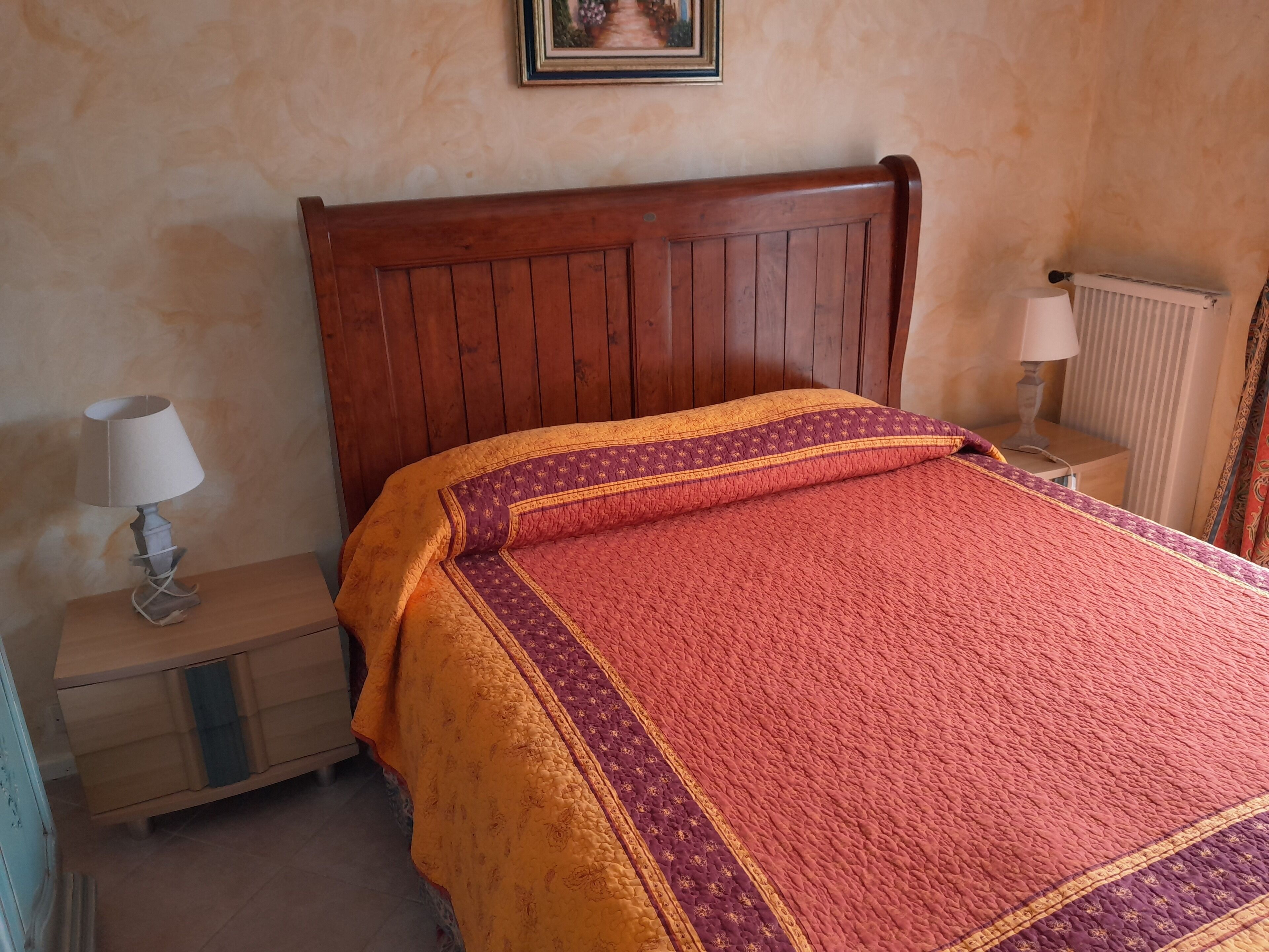 A bedroom with a wooden headboard, a bed with a patterned blanket, a nightstand with a lamp, and a radiator.