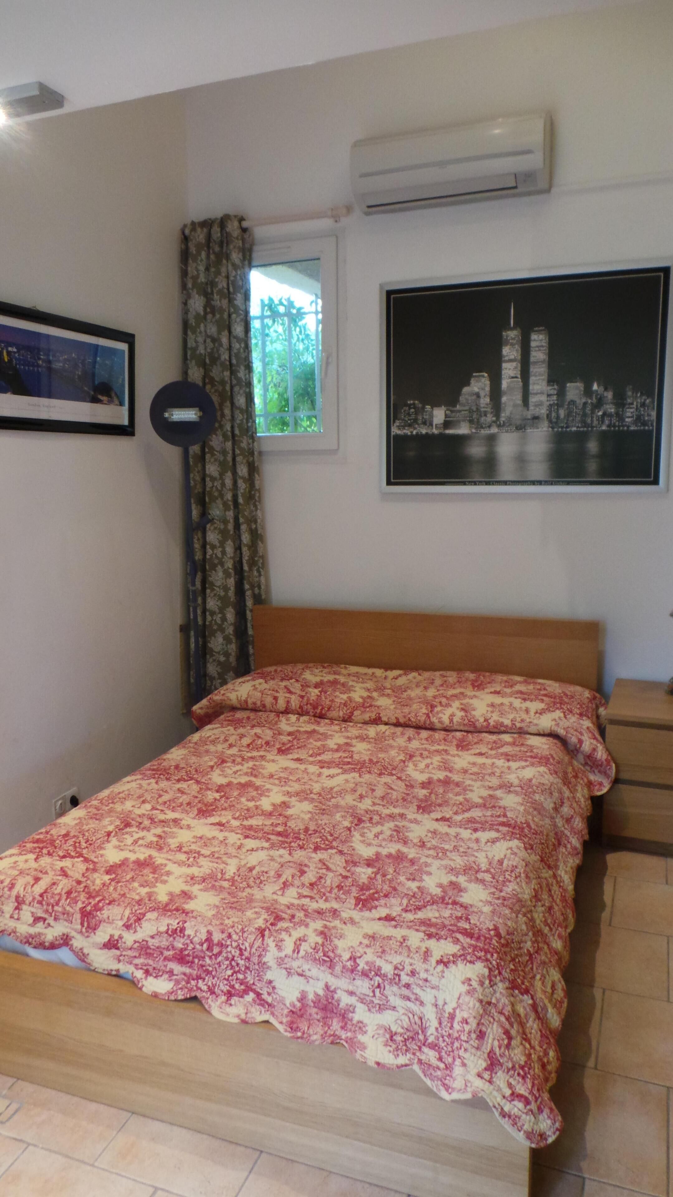 A bedroom with a bed, a window with curtains, an air conditioner, and a framed picture on the wall.