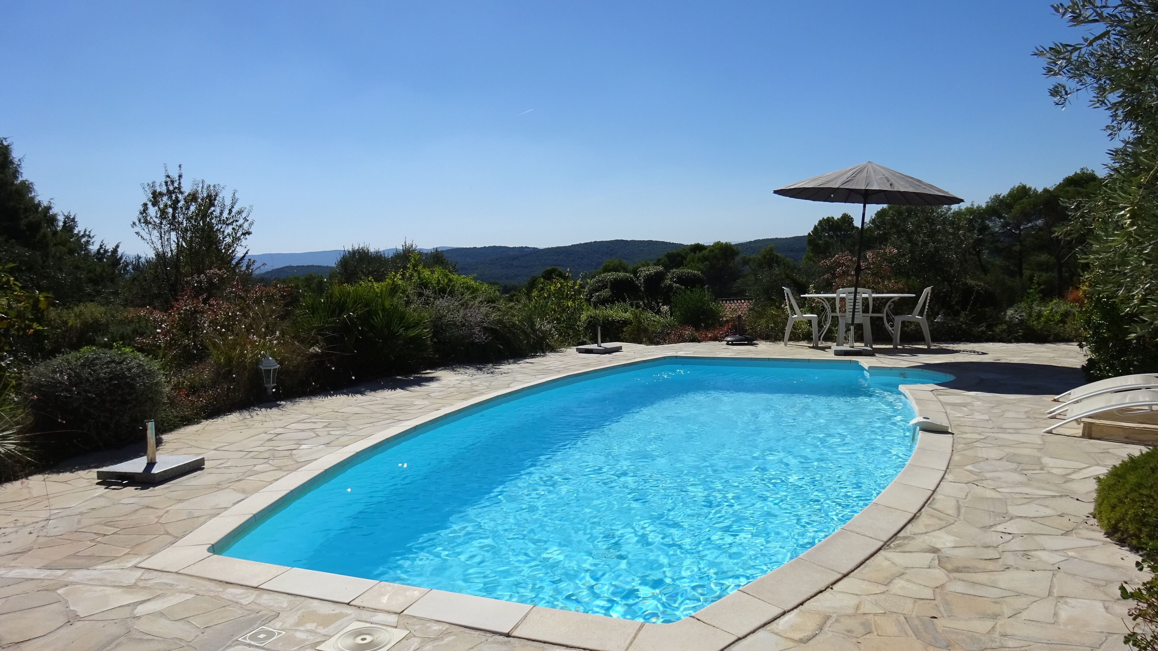 Ein rechteckiger Swimmingpool mit Steinbordierung, umgeben von einer Terrasse mit Steinplatten. Im Hintergrund stehen ein Tisch und Stühle mit Sonnenschirm.