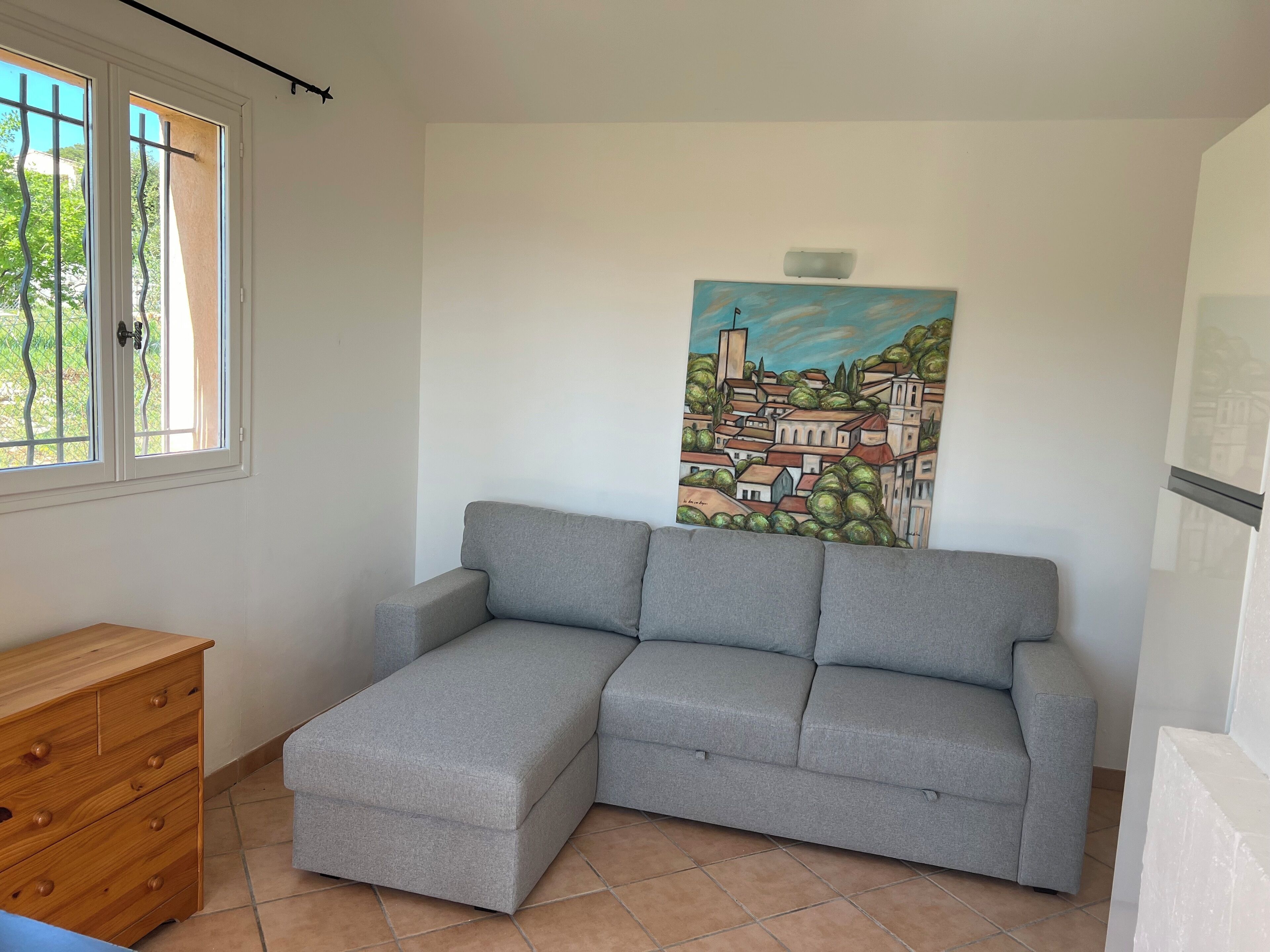 A room with a grey sofa, a wooden dresser, and a painting of a town on the wall.