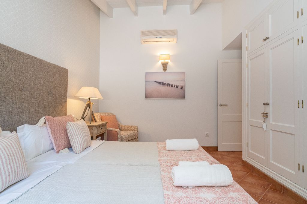 A bedroom with a bed, bedside lamps, a chair, a wardrobe, and a wall-mounted air conditioner.