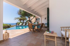 A covered outdoor area with a pool, wooden furniture, and a view of the sea.