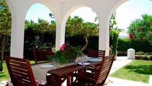 Un espace de restauration en plein air avec une table et des chaises en bois, entouré d’arches blanches et d’une végétation luxuriante.
