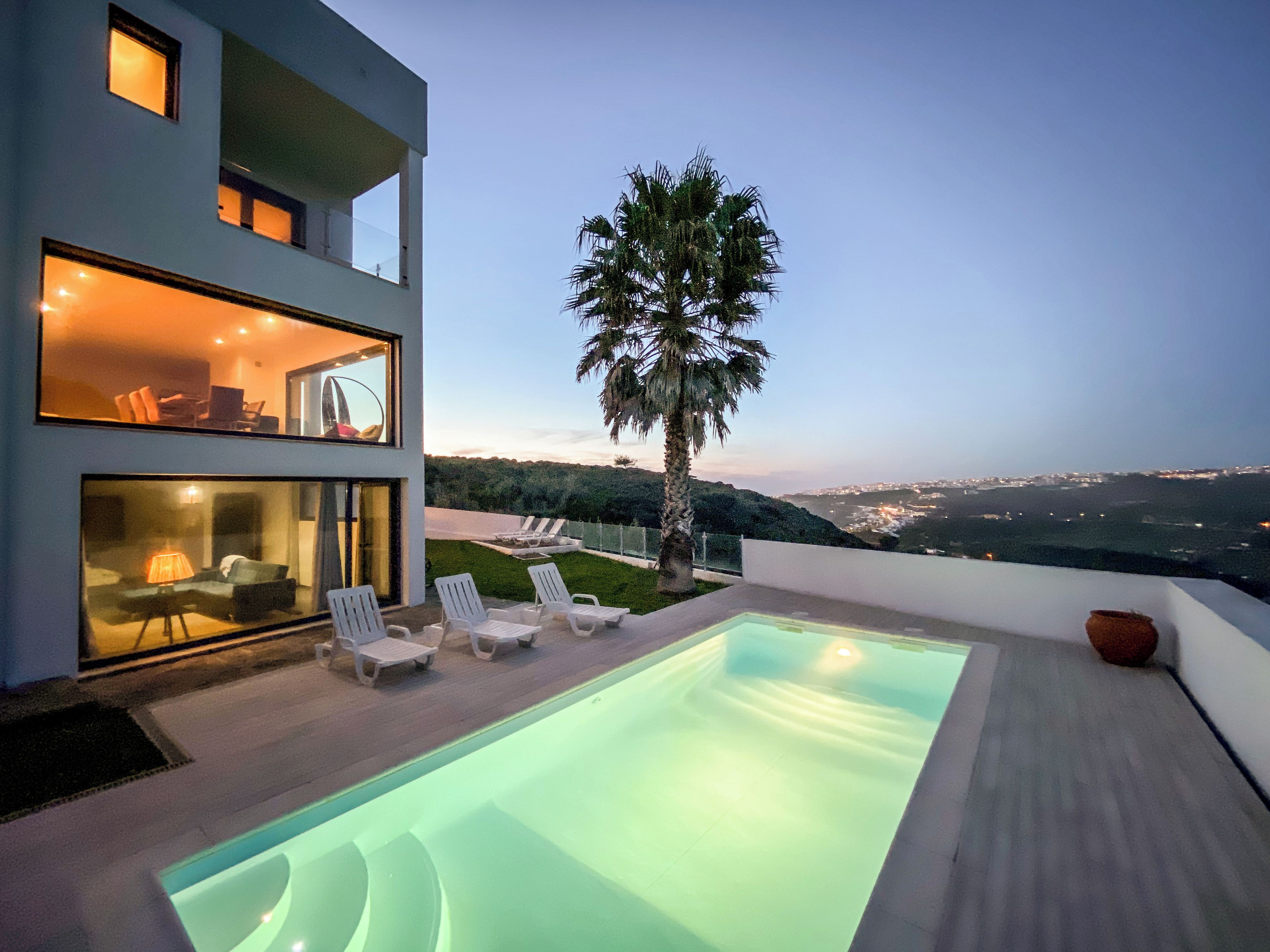 A modern building with a swimming pool, lounge chairs, and a view of the city at dusk.
