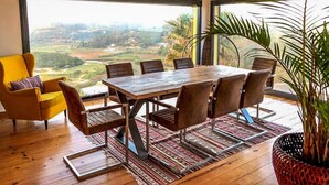 Un espace repas comprenant une grande table et des chaises, un fauteuil jaune et une vue sur un paysage grâce à de grandes fenêtres.