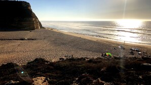 Une plage bordée d’une falaise rocheuse, un ciel dégagé et le soleil se couchant sur l’océan.