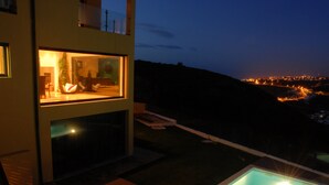 Une maison moderne dotée d’une piscine, d’une pièce entièrement vitrée et offrant une vue sur le paysage urbain la nuit.