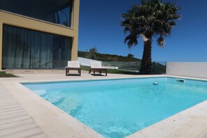 A swimming pool with two lounge chairs, a palm tree, and a modern building with large windows. - 5 Bedroom Villa  near Ericeira with sun terraces and heated outdoor pool (Ericiera)