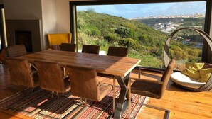 Un coin repas avec une table et des chaises en bois, une grande fenêtre donnant sur un paysage vallonné, et un fauteuil en osier avec un coussin.