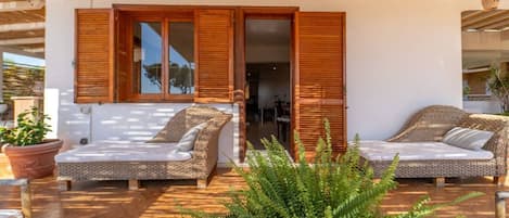 A covered outdoor seating area with wicker furniture and wooden shutters, a potted plant, and a view into the interior.