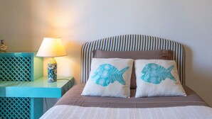 Ein Bett mit gestreifter Kopfteil, zwei Kissen mit Fischmotiven, eine Lampe und ein kleiner Tisch mit einer Dekoration.