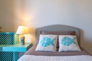 Ein Bett mit gestreifter Kopfteil, zwei Kissen mit Fischmotiven, eine Lampe und ein kleiner Tisch mit einer Dekoration.