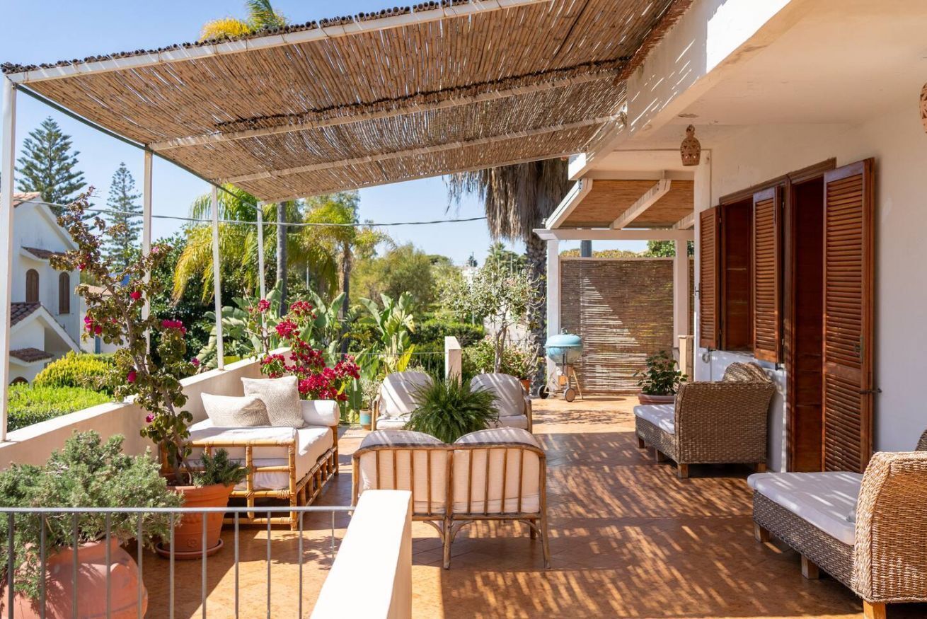 A covered outdoor seating area with wicker furniture, potted plants, and a view of the surrounding landscape.