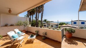 Ein Balkon mit Stühlen und Blumentöpfen, der auf eine Küstenlandschaft mit Palmen und Gebäuden blickt.