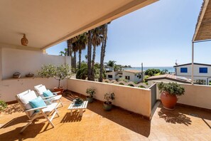 Ein Balkon mit Stühlen und Blumentöpfen, der auf eine Küstenlandschaft mit Palmen und Gebäuden blickt.