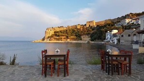 A seaside terrace with stone flooring, two wooden tables, and chairs, overlooking a calm sea and a historic cliff with buildings. - Elia apartment Holiday paradise next to the beach (Agia Triada, Koroni, Messinia)