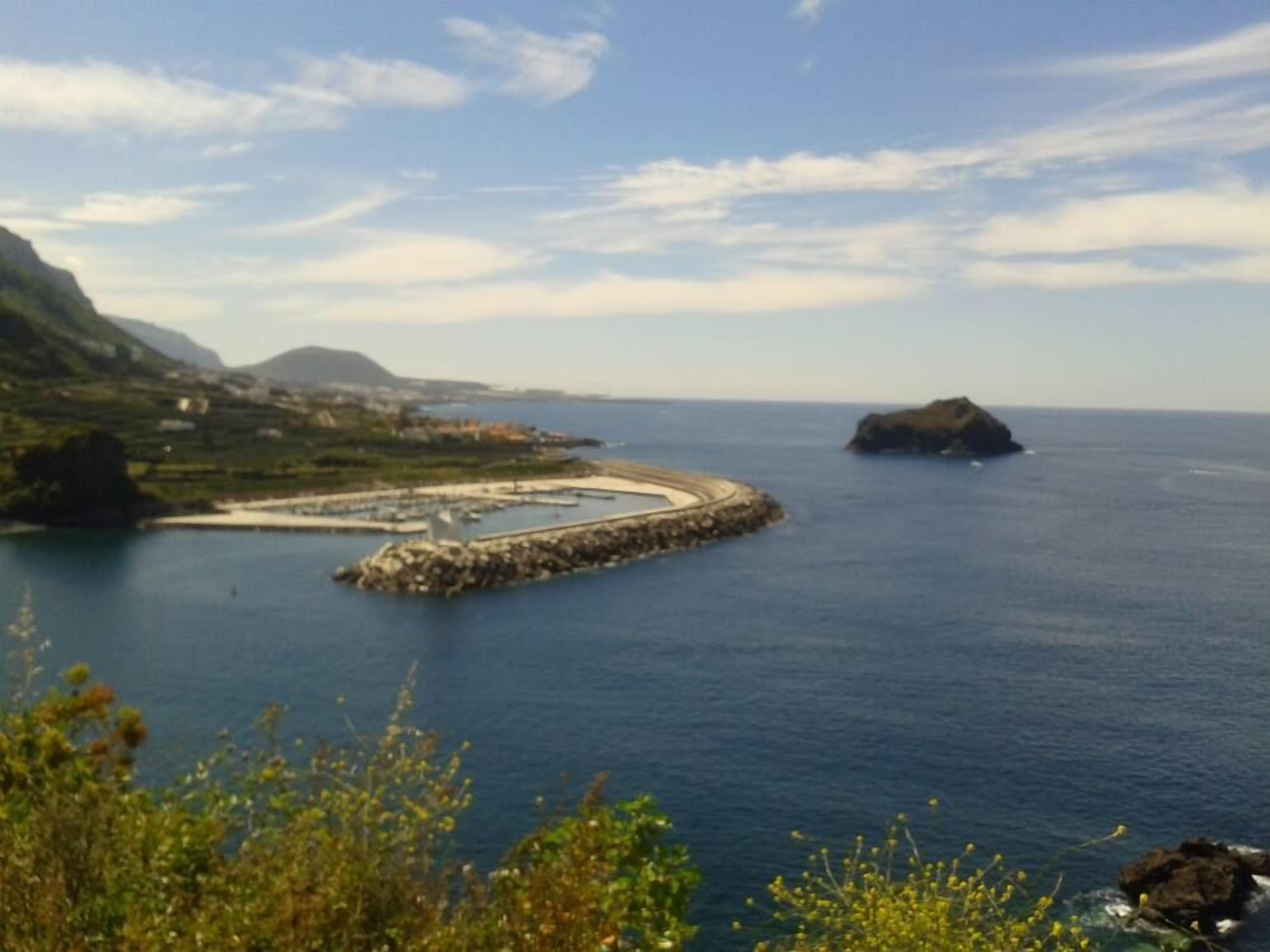 A coastal landscape with a circular stone structure surrounded by water, a distant island, and mountainous terrain.