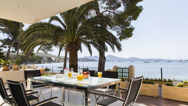 Une table à plateau en verre, quatre chaises, un saladier de fruits et une carafe de jus d’orange sont disposés sur une terrasse donnant sur un plan d’eau où se trouvent des bateaux.