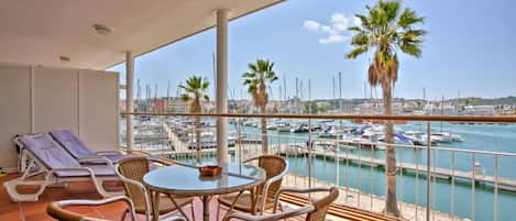Un balcón con mobiliario de mimbre, una mesa de vidrio y vistas a un puerto deportivo con barcos y palmeras.
