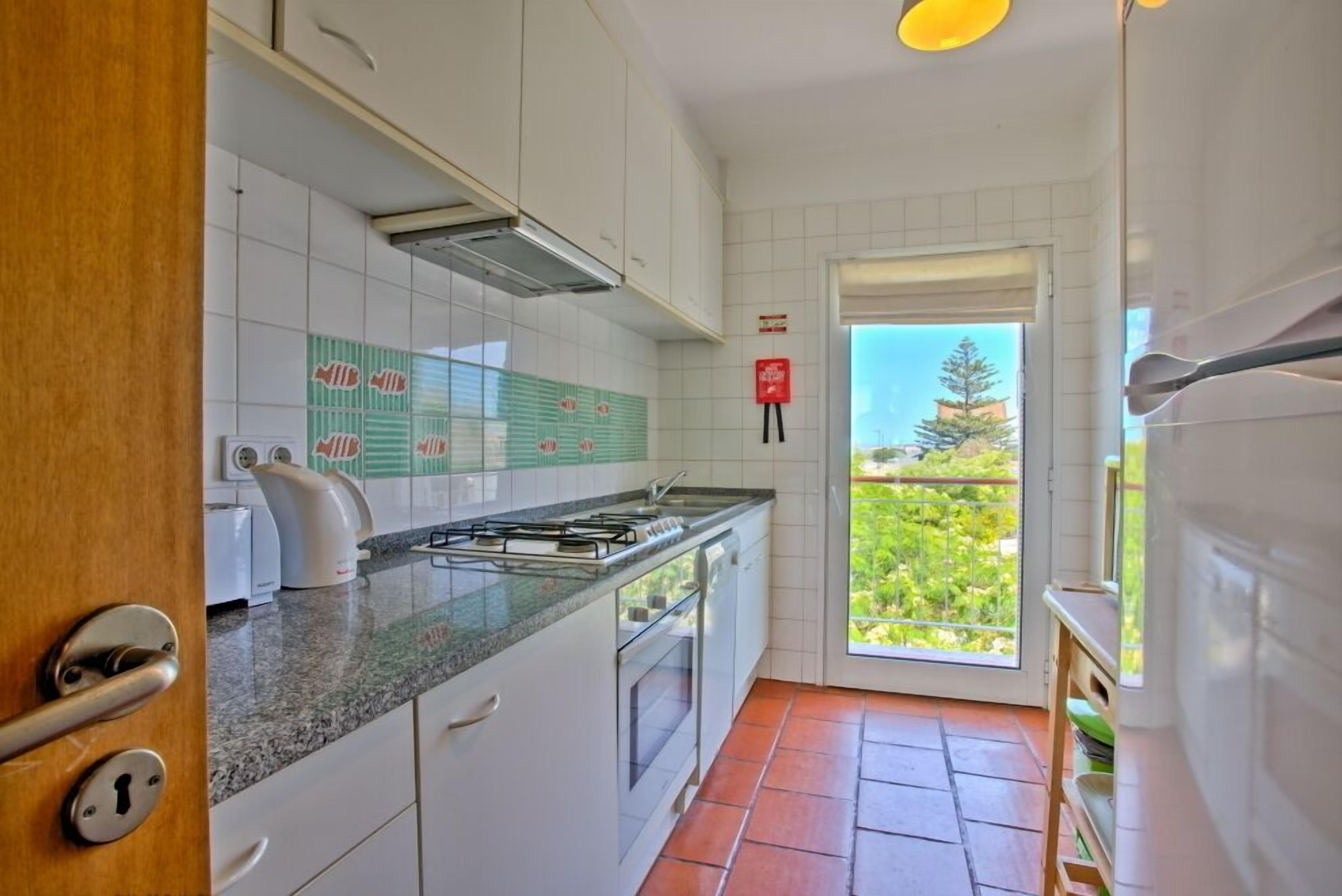 Une cuisine avec des meubles blancs, une cuisinière à gaz et une fenêtre donnant sur un paysage verdoyant.