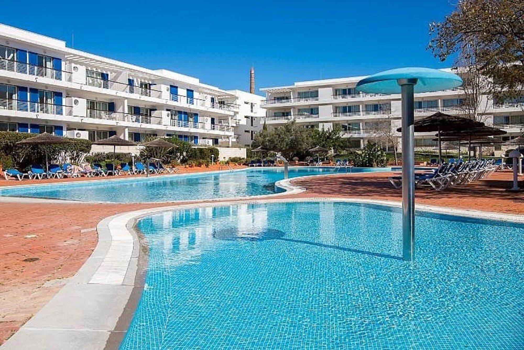 Uma piscina com espreguiçadeiras e guarda-sóis, cercada por um edifício moderno com várias varandas.