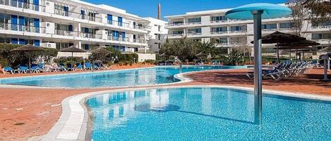 Uma piscina com espreguiçadeiras e guarda-sóis, cercada por um edifício moderno com várias varandas.
